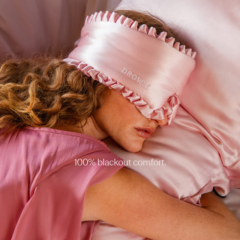 A woman with wavy hair lies on a bed wearing drowsysleepco’s Sleep and the City Powder Pink 100% mulberry silk sleep mask, matching pajamas, and resting on a pink silk pillowcase. Text reads 