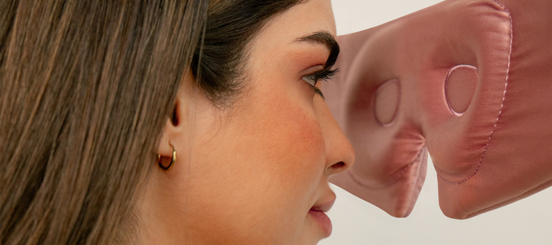 A woman with straight brown hair and a gold hoop earring is shown in profile, wearing a pink silk eye mask pushed up on her forehead. The background is neutral.