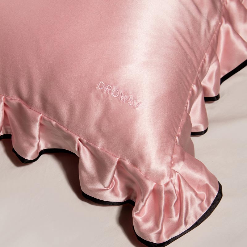 The Sleep and the City - Pink & Black Pillowcase by drowsysleepco, featuring shiny pink satin with ruffled edges and "DROWSY" embroidered, is shown resting on a light-colored surface.