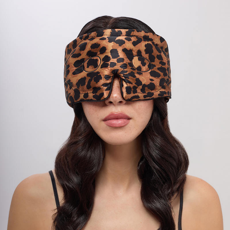A woman with long wavy hair wears the us.drowsysleepco Hotel Paradiso Wild Thing Eyelash Protecting Mask, a wide brown and black leopard-print blackout mask, covering her eyes and most of her face against a plain light background.