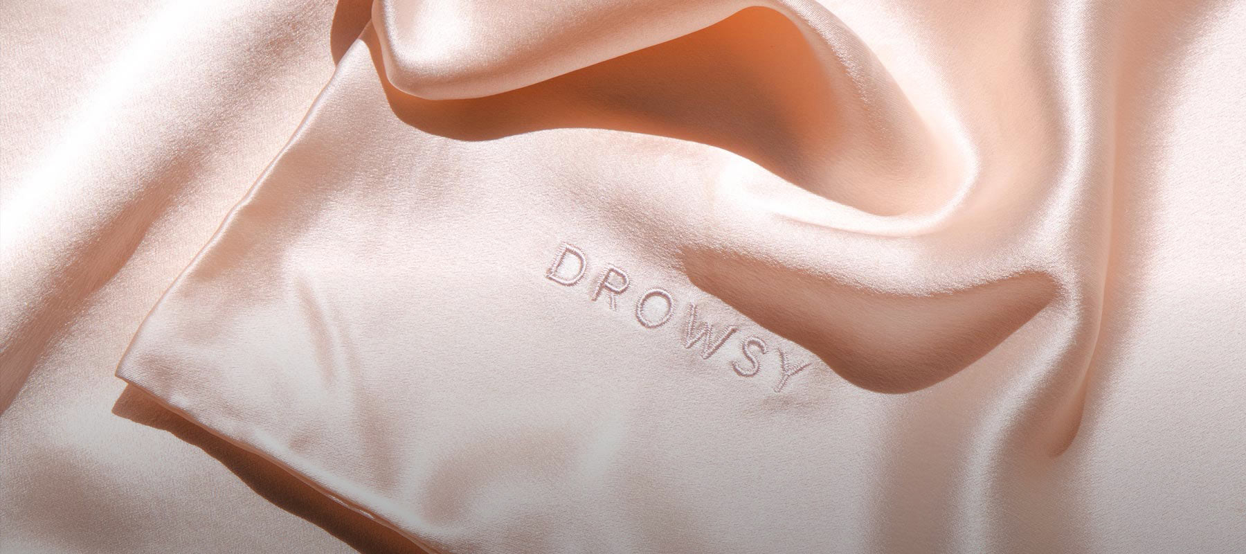 A light peach-colored silk fabric with the word DROWSY embroidered near the edge, softly folded and reflecting gentle light.