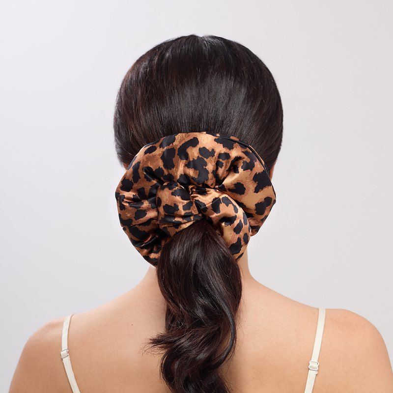 A woman with voluminous dark wavy hair, styled in a low ponytail and secured by the us.drowsysleepco Hotel Paradiso - Wild Thing Padded Scrunchie, wears a thin-strapped light top and stands against a plain background.