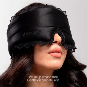 A woman wears the Paris Love Story - Black Jade Sleep Mask by drowsysleepco, crafted from black silk with lace trim and “DROWSY” embroidered on it. Text below: Wake up crease-free. Gentle on delicate skin for true beauty sleep.