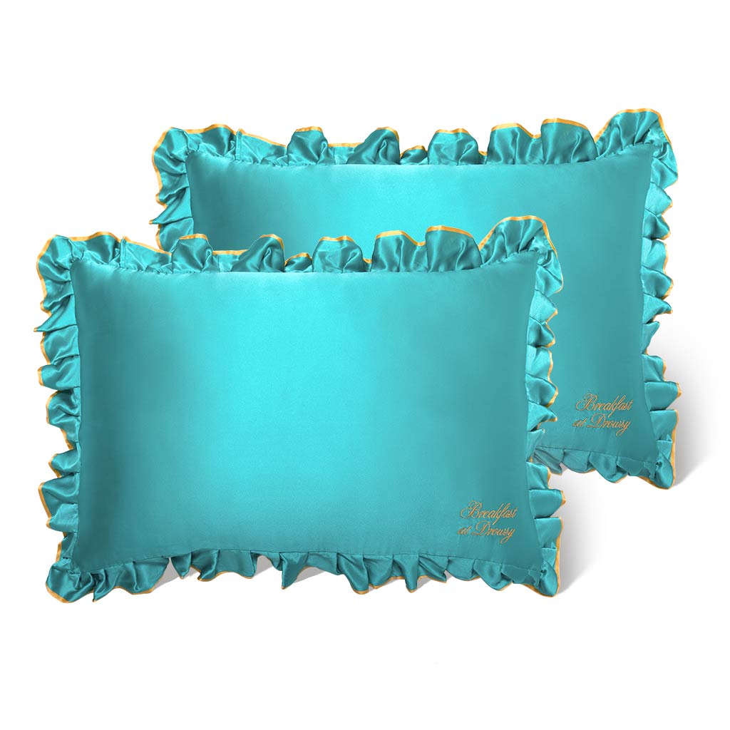 Two rectangular Breakfast at Drowsy - Turquoise Pillowcases by drowsysleepco feature ruffled edges, gold trim, and hair-protecting satin. Both pillowcases overlap slightly and display gold embroidery in the bottom right corner.
