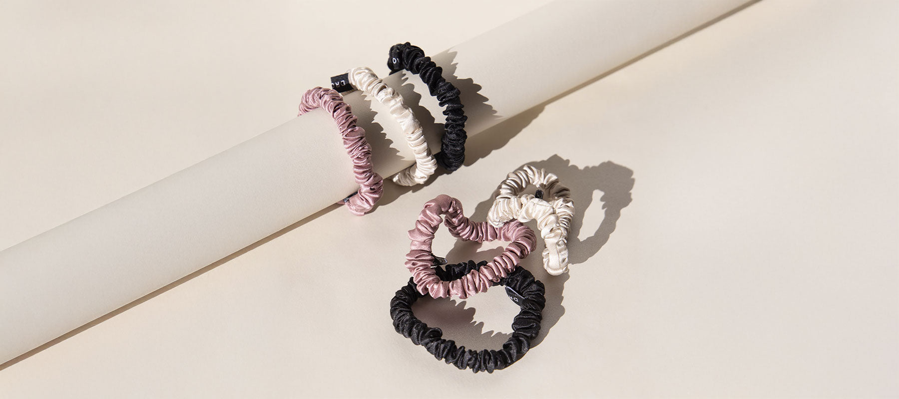 Three ruched fabric hair scrunchies in black, cream, and pink are looped around a cream-colored cylindrical rod, casting soft shadows on a beige background.