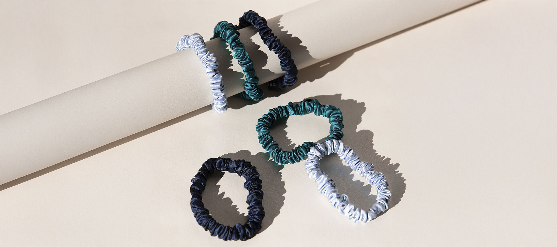 Three dark blue, two green, and two light blue fabric scrunchies are displayed on and beside a beige cylindrical object, casting soft shadows on a light beige surface.