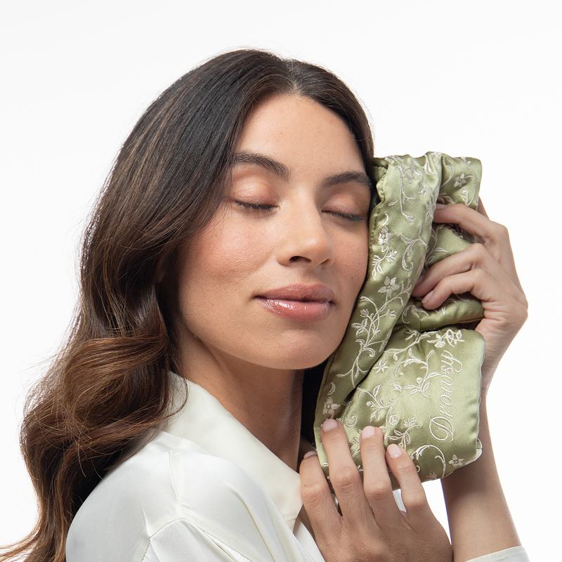A person with eyes closed holds a sage green Pride & Prejudice - Sage Eyelash Protecting Sleep Mask with floral embroidery against their cheek.