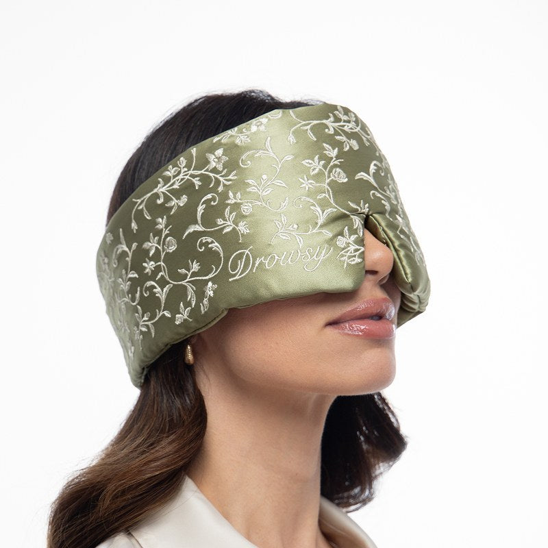 A person wearing a sage green Pride & Prejudice - Sage Eyelash Protecting Sleep Mask embroidered with white floral vines and the word Drowsy.
