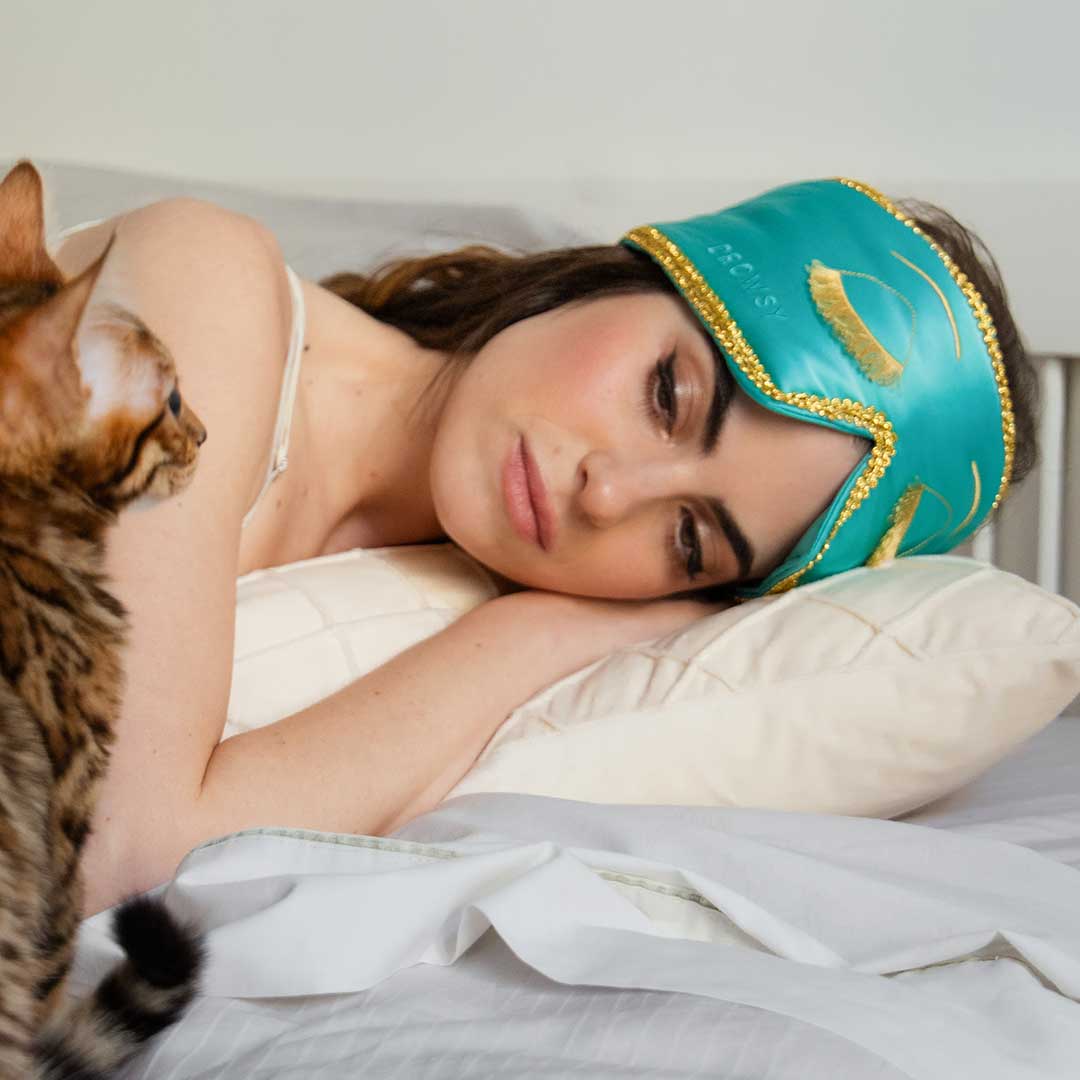 A woman wearing a drowsysleepco 2-Pack Sleep Mask | Breakfast at Drowsy in turquoise with gold details rests on her pillow and looks at a brown tabby cat nearby.