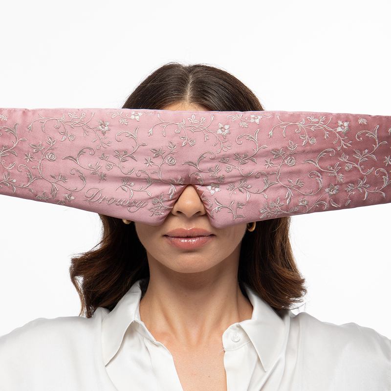 A person wearing a dusty pink mulberry silk Pride & Prejudice - Sage + English Rose Eyelash Protecting Mask embroidered with silver floral patterns.