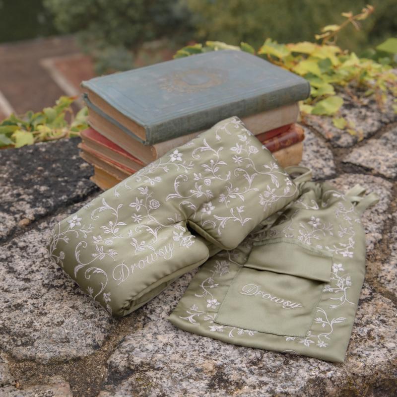 A sage green Pride & Prejudice - Sage Sleep Mask + Pouch in mulberry silk with a white floral vine pattern, resting with its matching drawstring pouch on a stack of antique books atop a stone wall.