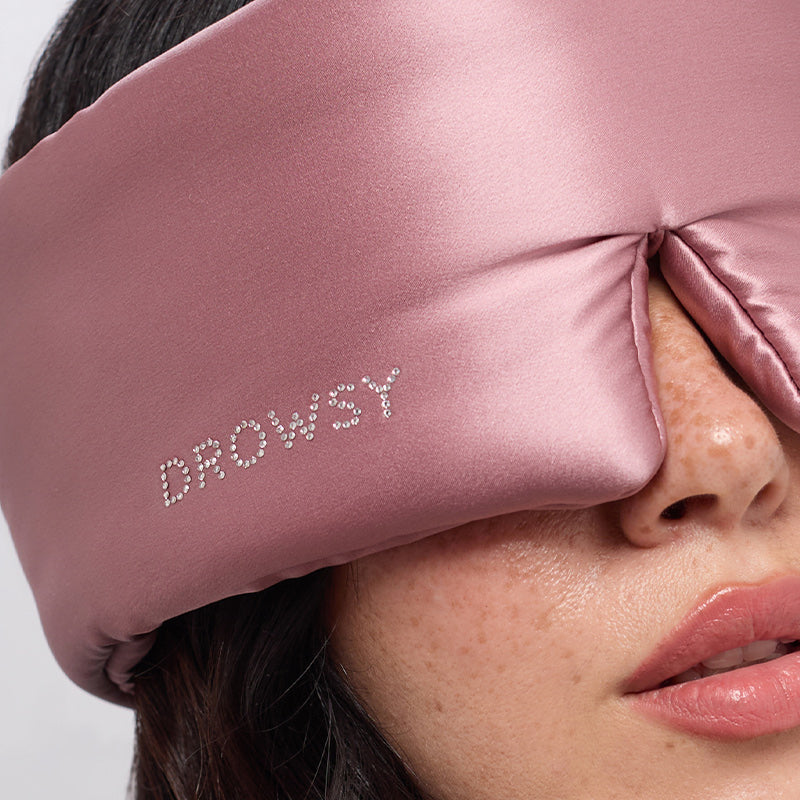 A close-up of a woman wearing the drowsysleepco Damask Rose sleep mask, featuring a Swarovski®-embellished logo, covering her eyes and part of her nose.