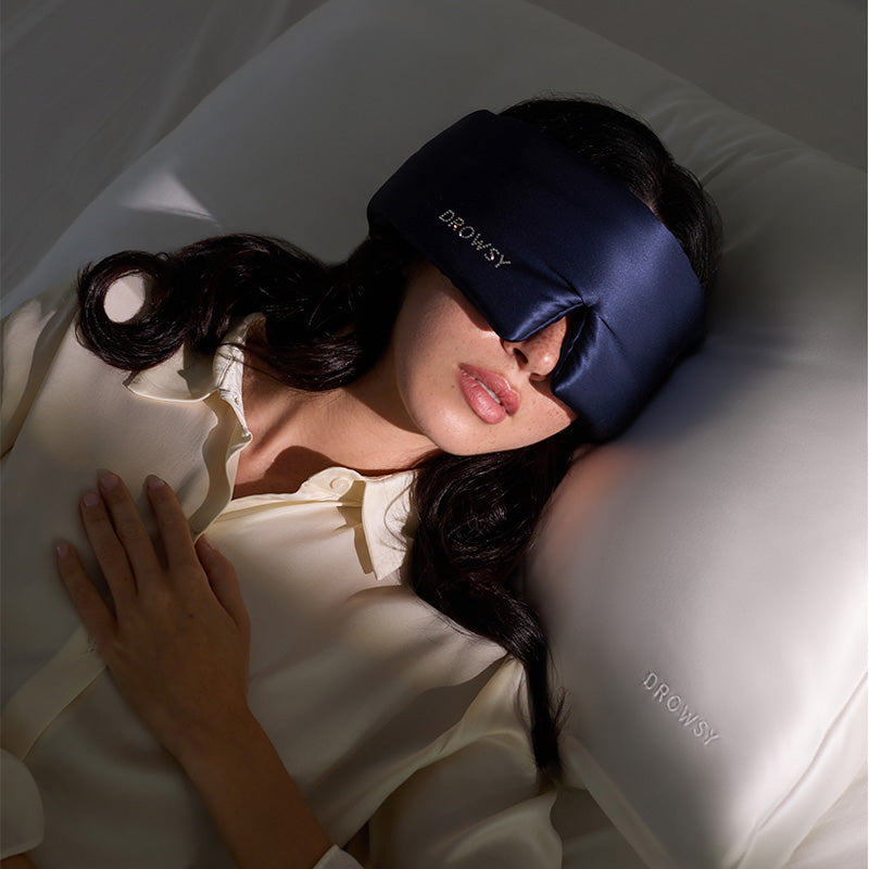A woman lies on her back in bed, wearing a drowsysleepco Midnight Blue sleep mask with a Swarovski® embellished logo. One hand rests on her chest and she uses a white pillow embroidered with 