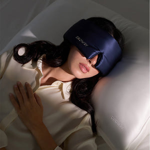 A woman lies on her back in bed, wearing a drowsysleepco Midnight Blue sleep mask with a Swarovski® embellished logo. One hand rests on her chest and she uses a white pillow embroidered with "DROWSY.